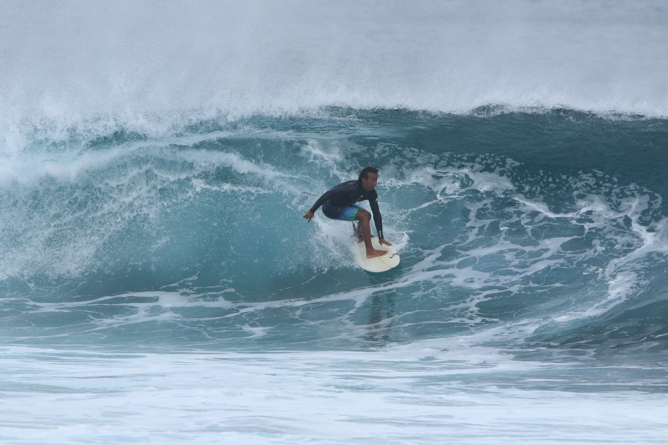 A surfer in a dark rash vest crouched low inside a steep, breaking blue wave.