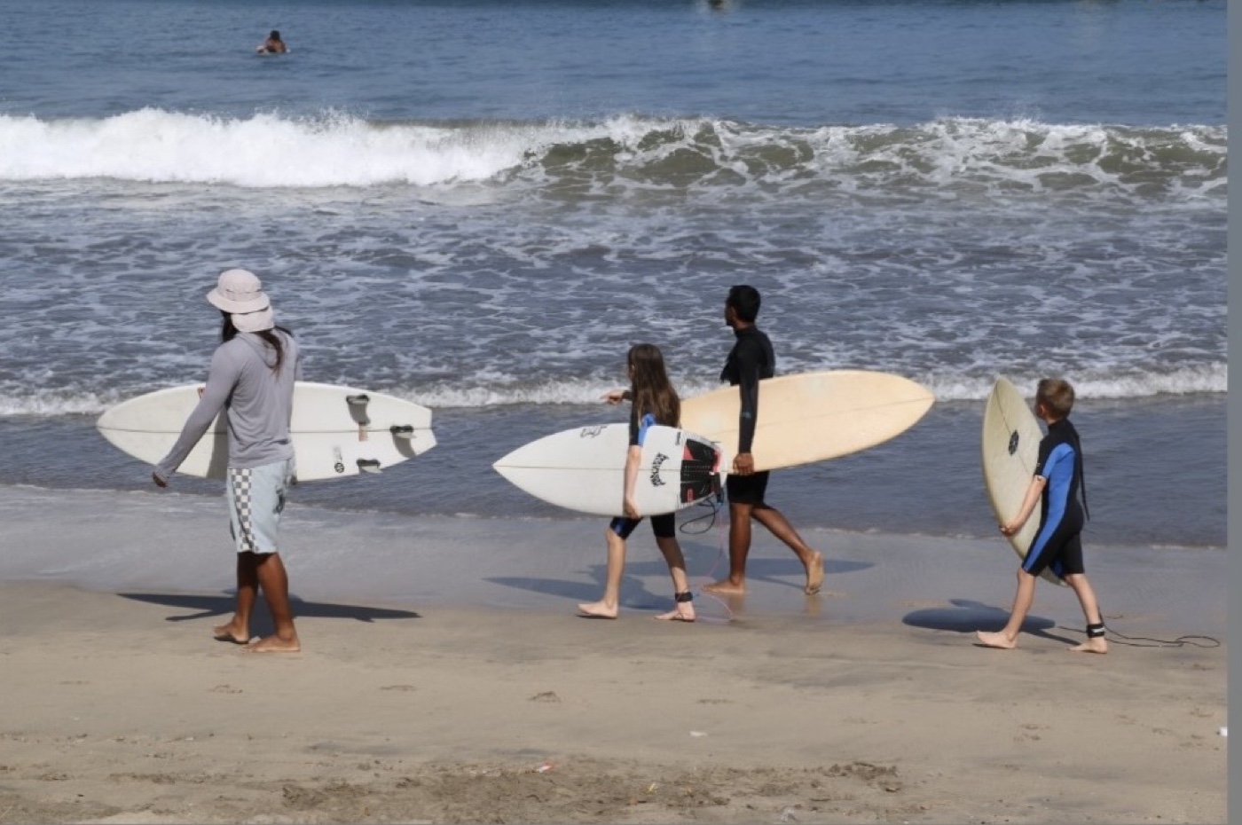 An instructor and three young students walking along the shoreline carrying surfboards.