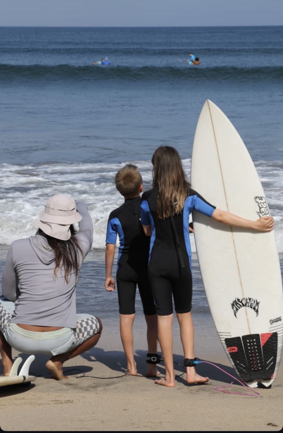 An instructor crouched on the sand with two children in wetsuits, all watching the waves.